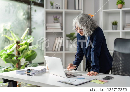 Elderly female business executive analyzing data on laptop while reviewing documents in a modern office Elderly female business executive analyzing data on laptop while reviewing documents in a modern office 122717276