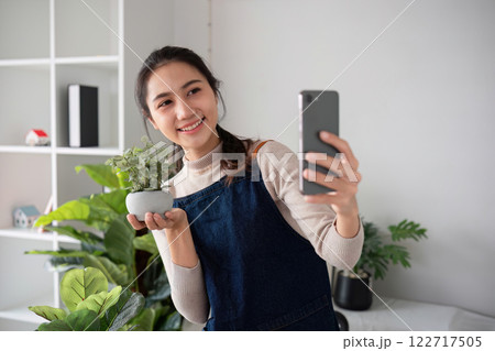 A young Asian woman uses her phone to take photos and enjoys planting plants in her garden to create a pleasant atmosphere in the house. 122717505