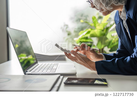 Elderly businesswoman reviewing financial data on a calculator and laptop in her stylish office Elderly businesswoman reviewing financial data on a calculator and laptop in her stylish office 122717730