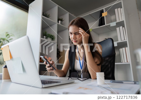 Stressed businesswoman managing work pressure while using smartphone in contemporary office Stressed businesswoman managing work pressure while using smartphone in contemporary office 122717836