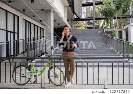 A young Asian businessman rides a bicycle to work. Standing outside the office talking on the phone and talking about business. Concept of reducing energy and reducing air pollution. 122717840