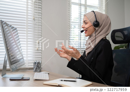 Call center worker, young Muslim woman wearing hijab, talking to customer on call phone on computer in customer service office Call center worker, young Muslim woman wearing hijab, talking to customer on call phone on computer in customer service office 122717959