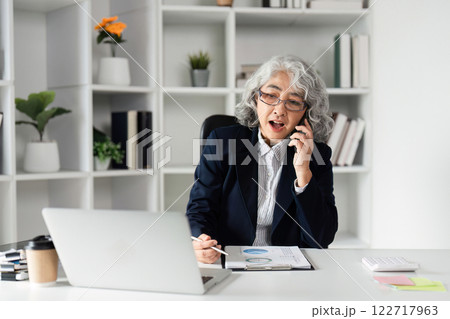 Elderly Businesswoman Engaged in a Phone Call at Her Office Desk, Managing Company Affairs with Confidence and Expertise 122717963