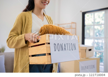 A cute Asian woman stands holding a box, sorting unwanted clothes for donation. A cute Asian woman stands holding a box, sorting unwanted clothes for donation. 122718004
