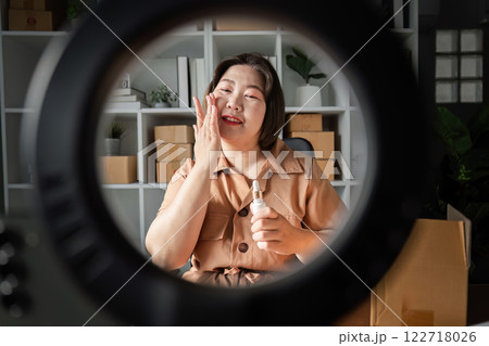 Young woman applying skincare cream while livestreaming sales from a modern home office, engaging viewers through a ring light Young woman applying skincare cream while livestreaming sales from a modern home office, engaging viewers through a ring light 122718026