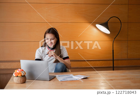 Young Woman Sitting Indoors Focused on Paperwork with Laptop and Lamp in Modern Room Setting Young Woman Sitting Indoors Focused on Paperwork with Laptop and Lamp in Modern Room Setting 122718043