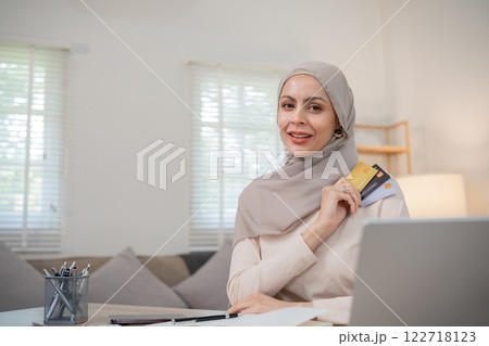 A young Muslim woman wearing a hijab sits contentedly shopping on her laptop, paying through an online banking app and holding a credit card. A young Muslim woman wearing a hijab sits contentedly shopping on her laptop, paying through an online banking app and holding a credit card. 122718123