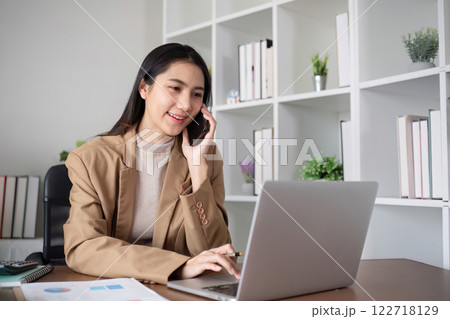 Young Asian business woman sits on the phone in an online business meeting using a laptop in a modern home office decorated with shady green plants. 122718129