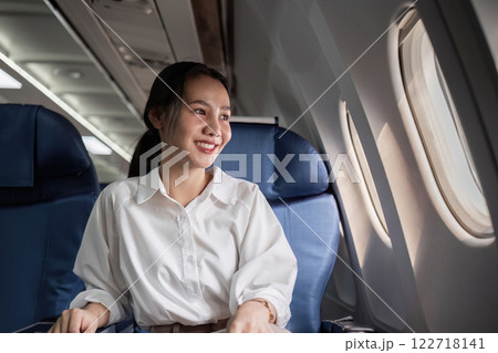 Confident Businesswoman Traveling by Plane, Seated in Comfortable Airplane Cabin, Looking Out Window with a Smile Confident Businesswoman Traveling by Plane, Seated in Comfortable Airplane Cabin, Looking Out Window with a Smile 122718141