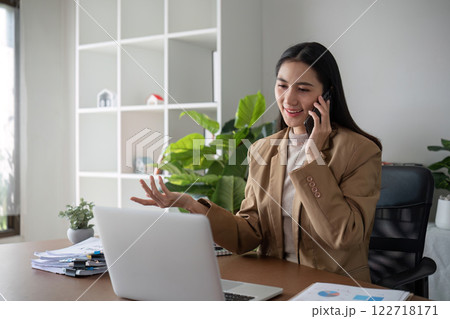 Young Asian business woman sits on the phone in an online business meeting using a laptop in a modern home office decorated with shady green plants. Young Asian business woman sits on the phone in an online business meeting using a laptop in a modern home office decorated with shady green plants. 122718171