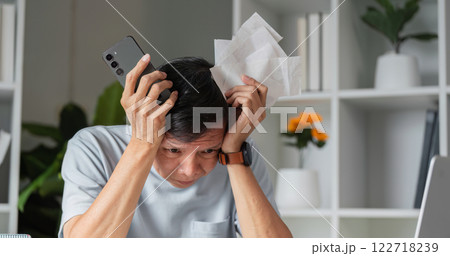Frustrated man dealing with financial problems and expenses while holding his smartphone and paperwork at home 122718239