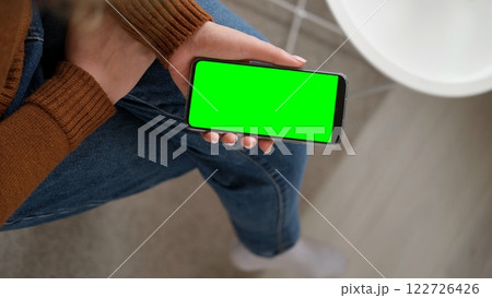 Young woman sitting at home holding smartphone green mock-up screen in hand. Female person use chroma key mobile phone. Horizontal mode. Touching, swipe display, tapping, surfing internet social media Young woman sitting at home holding smartphone green mock-up screen in hand. Female person use chroma key mobile phone. Horizontal mode. Touching, swipe display, tapping, surfing internet social media 122726426