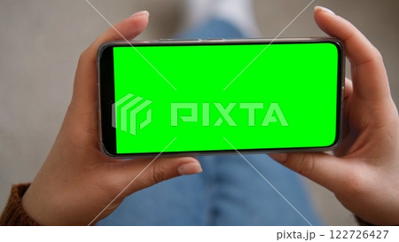 Young woman sitting at home holding smartphone green mock-up screen in hand. Female person use chroma key mobile phone. Horizontal mode. Touching, swipe display, tapping, surfing internet social media 122726427