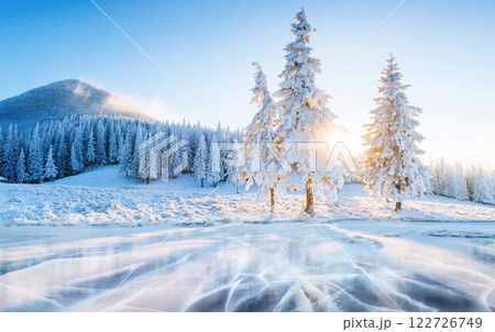 Blue ice and cracks on the surface of the ice. Frozen lake under a blue sky in the winter. Beautiful dawn, rays of the sun through the branches. Carpathian, Ukraine, Europe 122726749