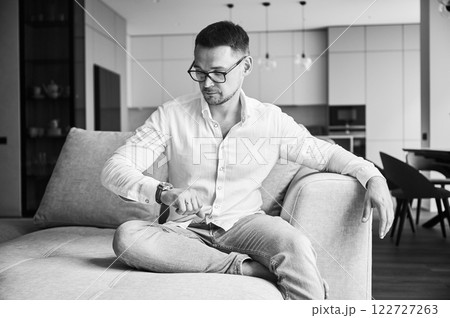Busy man checking time on his watch. Young man waiting for important meeting. Handsome male in casual clothes sitting on a couch. Black and white image. Busy man checking time on his watch. Young man waiting for important meeting. Handsome male in casual clothes sitting on a couch. Black and white image. 122727263