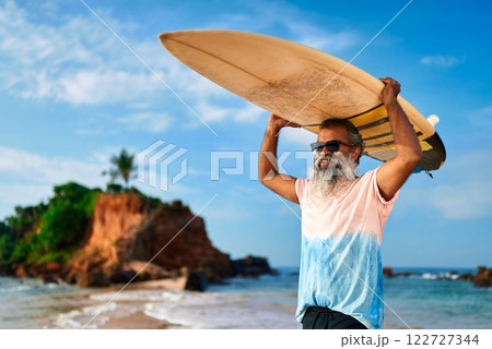 Senior ethnic man with surfboard on tropical beach. Grey-haired male surfer in sunglasses holds board over head. Active lifestyle older adults. Ocean waves under sunny blue sky. Retirement adventures. Senior ethnic man with surfboard on tropical beach. Grey-haired male surfer in sunglasses holds board over head. Active lifestyle older adults. Ocean waves under sunny blue sky. Retirement adventures. 122727344
