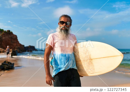 Older ethnic man on sunny beach with surfboard. Senior male, casual surf gear. Active lifestyle, enjoys sea, surf culture. Fit elderly, vibrant mood. Ocean waves, surf beach. Mature surfer. Older ethnic man on sunny beach with surfboard. Senior male, casual surf gear. Active lifestyle, enjoys sea, surf culture. Fit elderly, vibrant mood. Ocean waves, surf beach. Mature surfer. 122727345