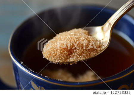 Person adding cane sugar with a spoon to cup of fresh aromatic coffee. Close up of teaspoon putting brown sugar into hot steamed drink mug. Sugar Being Poured into Cup of Coffee Americano, Close-Up 122728323