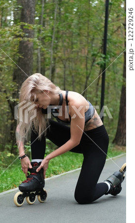 Cute blonde puts on roller skates. She feels free and light while roller skating alone in the city park. Stunning Caucasian woman with a beautiful figure uses roller skates for outdoor sports. 122729246