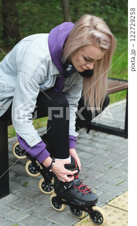 Cute blonde puts on roller skates. She feels free and light while roller skating alone in the city park. Stunning Caucasian woman with a beautiful figure uses roller skates for outdoor sports. Cute blonde puts on roller skates. She feels free and light while roller skating alone in the city park. Stunning Caucasian woman with a beautiful figure uses roller skates for outdoor sports. 122729258