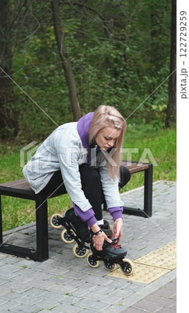 Cute blonde puts on roller skates. She feels free and light while roller skating alone in the city park. Stunning Caucasian woman with a beautiful figure uses roller skates for outdoor sports. Cute blonde puts on roller skates. She feels free and light while roller skating alone in the city park. Stunning Caucasian woman with a beautiful figure uses roller skates for outdoor sports. 122729259