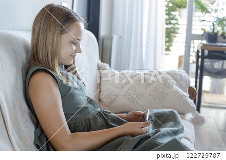 Smiling young girl using the phone, playing the game, sitting on sofa at cozy home.  122729787