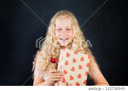 Beautiful young girl holding gift box with red heart. Blond child with long healthy curly hair Beautiful young girl holding gift box with red heart. Blond child with long healthy curly hair 122730543