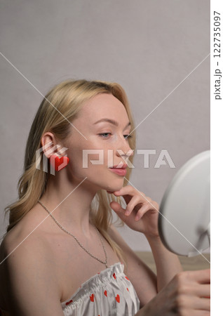 Blonde smiling girl in a white pajama heart pattern with red heart shape earring look at round mirror near wooden table. White wall background. Valentine's Day preparation Blonde smiling girl in a white pajama heart pattern with red heart shape earring look at round mirror near wooden table. White wall background. Valentine's Day preparation 122735097