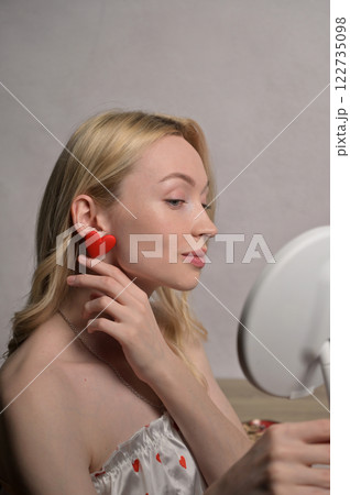 Blonde smiling girl in a white pajama heart pattern touching a red heart shape earring look at round mirror near wooden table. White wall background. Valentine's Day preparation 122735098
