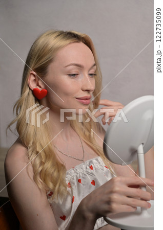 Blonde smiling girl in a white pajama heart pattern with red heart shape earring touching hair and look at round mirror near wooden table. White wall background. Valentine's Day preparation 122735099