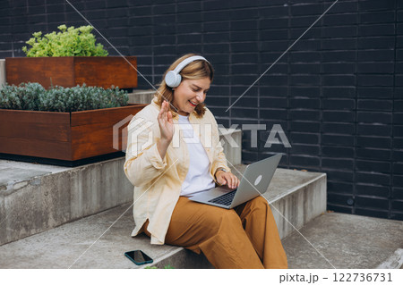 Happy 30s woman in headphones doing business on laptop and smiles. Young woman enjoy her work outdoor. Remote work, freelance. Young pretty female talking on video call and waving hand on stairs Happy 30s woman in headphones doing business on laptop and smiles. Young woman enjoy her work outdoor. Remote work, freelance. Young pretty female talking on video call and waving hand on stairs 122736731