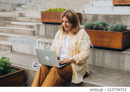 Smiling woman sitting on stairs working on laptop, preparing for exams outdoors. Technology, education and remote working concept 122736732
