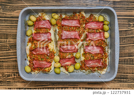 Roasted fish fillets are layered with bacon and herb sauce, accompanied by green olives 122737931