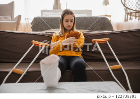 Young girl with broken leg cast sitting on sofa. Sad little kid hugging her teddy bear recovering from broken leg at home. Girl using cast and crutches. Accident injury fracture treatment foot bandage Young girl with broken leg cast sitting on sofa. Sad little kid hugging her teddy bear recovering from broken leg at home. Girl using cast and crutches. Accident injury fracture treatment foot bandage 122738293
