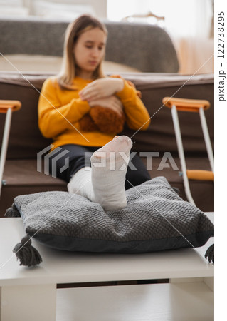 Broken leg. Young girl with broken leg cast sitting on sofa. Sad little kid recovering from broken leg at home. Girl using cast and crutches for rehabilitation. Accident injury fracture treatment 122738295