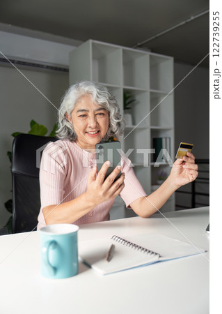 Happy elderly woman shopping online with credit card and smartphone in her stylish home office Happy elderly woman shopping online with credit card and smartphone in her stylish home office 122739255