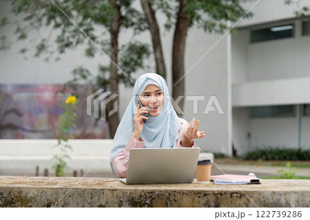 Muslim businesswoman in hijab talking on the phone while working on a laptop outdoors. 122739286