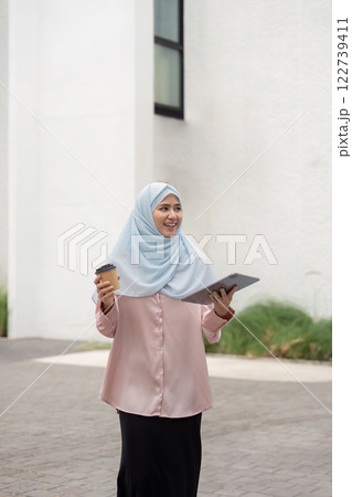 Muslim woman holding coffee and tablet while walking in a business environment. 122739411