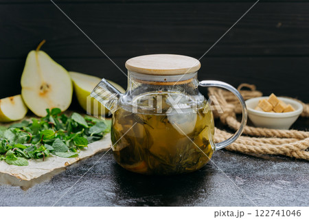 Pear and mint herbal tea in cup with glass teapot, fresh ingredients, and sugar cubes on dark table 122741046
