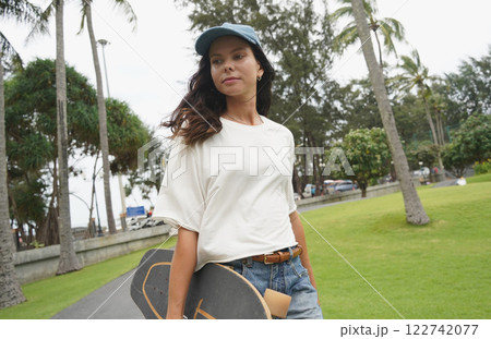 A woman is walking at the city park while carrying a skateboard in her hands A woman is walking at the city park while carrying a skateboard in her hands 122742077