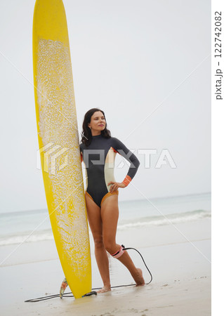 A woman in a stylish wet suit confidently holds a surfboard on the beach 122742082