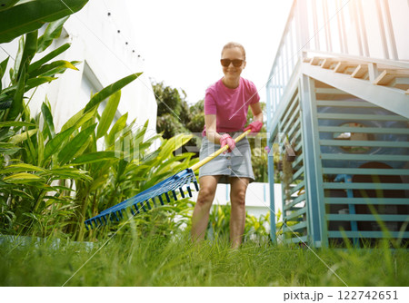 A woman raking the lawn at the backyard of her house  122742651