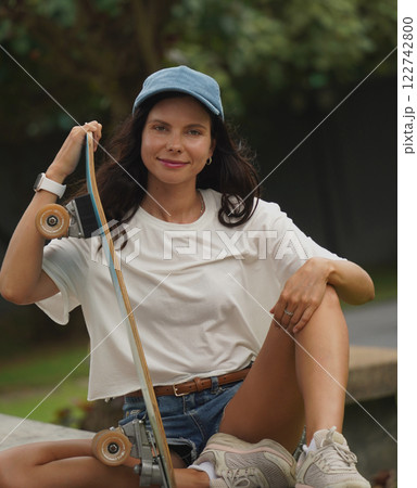 A young woman sits comfortably on a low wall at the park holding a skateboard in her hands 122742800