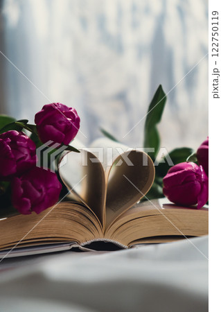 Book pages folded into heart with pink fresh tulips on white bed with trendy deep shadows. Spring flowers gift present. Concept of love literature and education Romantic date postcard wallpaper design 122750119