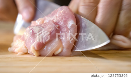A chef using a kitchen knife slices uncooked chicken meat on a chopping board, the close up view of preparing food ingredients. A chef using a kitchen knife slices uncooked chicken meat on a chopping board, the close up view of preparing food ingredients. 122753893