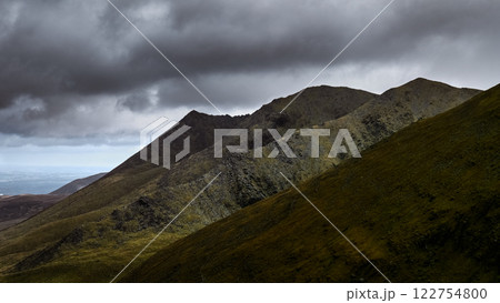 Jagged mountain ridges under a dramatic, cloudy sky, visible lower terrain with some greenery. 122754800