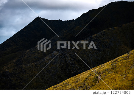 Multiple dark mountain summits, layered and steep, with a stormy sky above and glimpses of lower land. 122754803