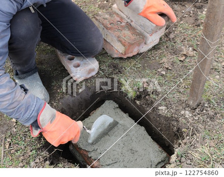 worker in orange gloves laying and leveling a layer of fresh cement with a trowel in a hole in the ground on a block with a string stretched nearby as a level, preparing for foundation work worker in orange gloves laying and leveling a layer of fresh cement with a trowel in a hole in the ground on a block with a string stretched nearby as a level, preparing for foundation work 122754860