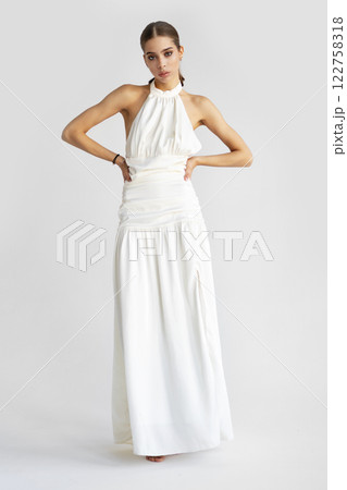 Young Beautiful Woman with dark hair stands and poses on isolated white background, wearing white maxi dress, looking in camera. Glamor fashion model. Beauty, skincare concept 122758318