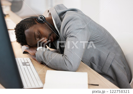 Tired CRM call center agent sleeping on computer desk at work office with headset on. Exhausted young business man taking a nap on table. African American male feeling burnout resting in workplace. 122758321